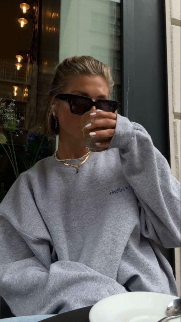 Woman in oversized gray sweatshirt with sunglasses enjoying a drink outdoors.