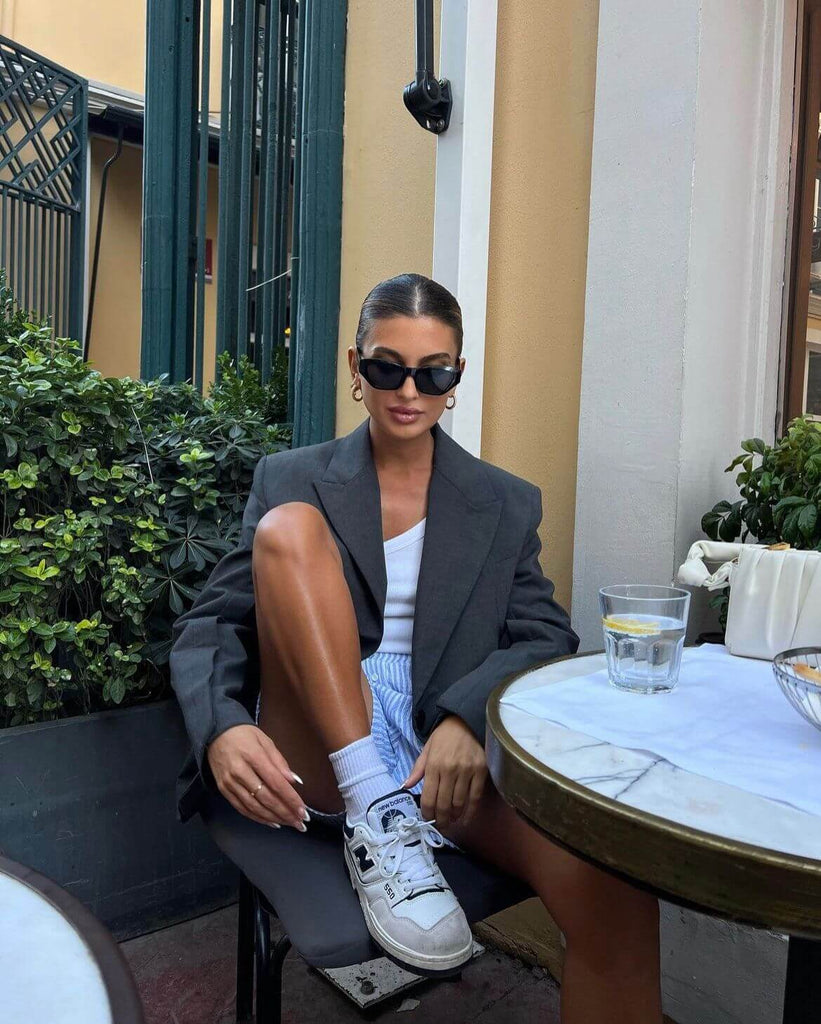 Stylish woman in oversized blazer and sunglasses, wearing sneakers at a cafe with greenery.