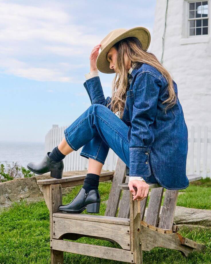 Stylish woman wearing ankle boots, denim outfit, and hat, seated on a wooden chair with a coastal background.