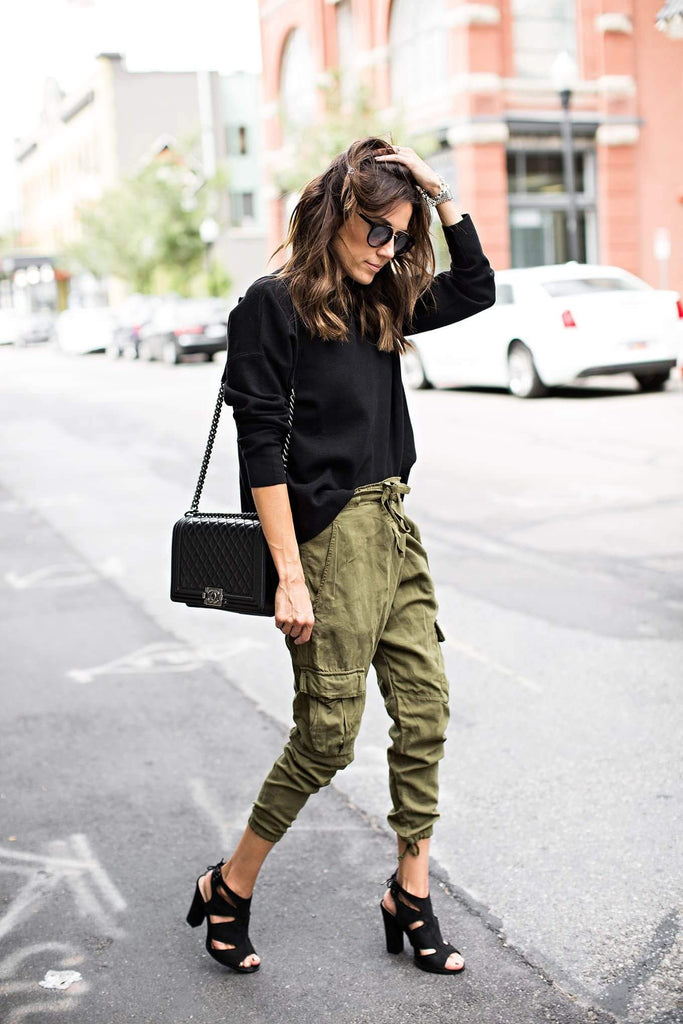 Stylish woman wearing black sweater and olive cargo pants, accessorized with a handbag and heels, walking on urban street.