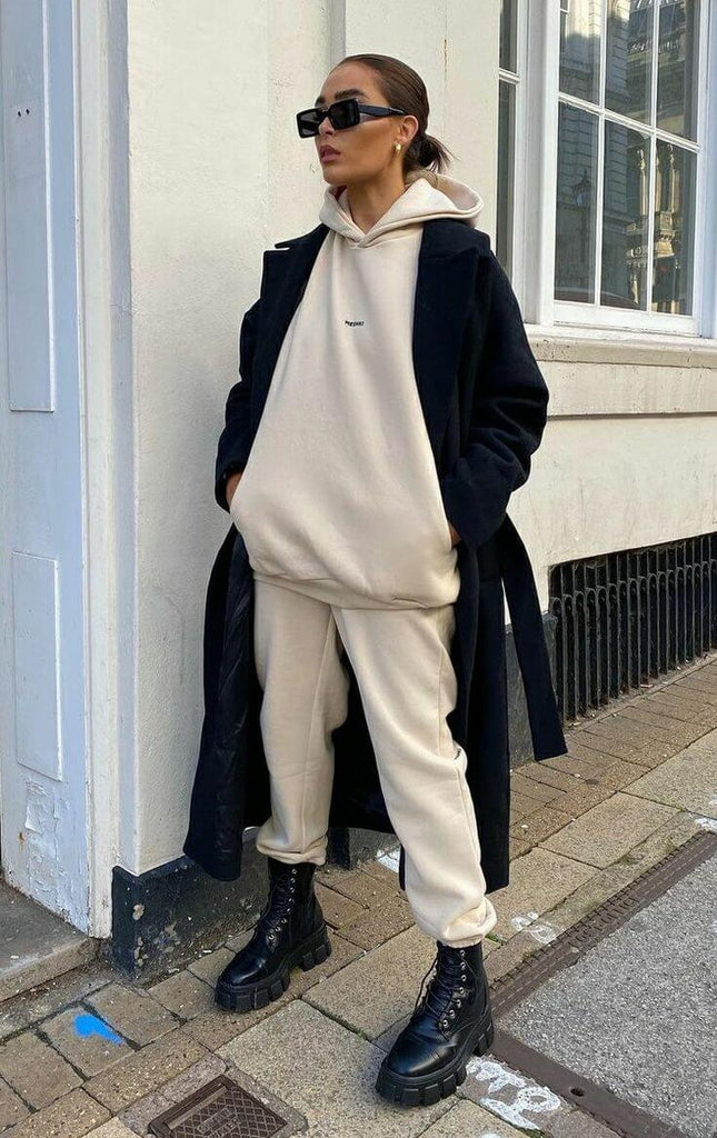 Stylish woman in a beige hoodie and matching pants, wearing a long black coat and sunglasses, showcasing casual fashion.